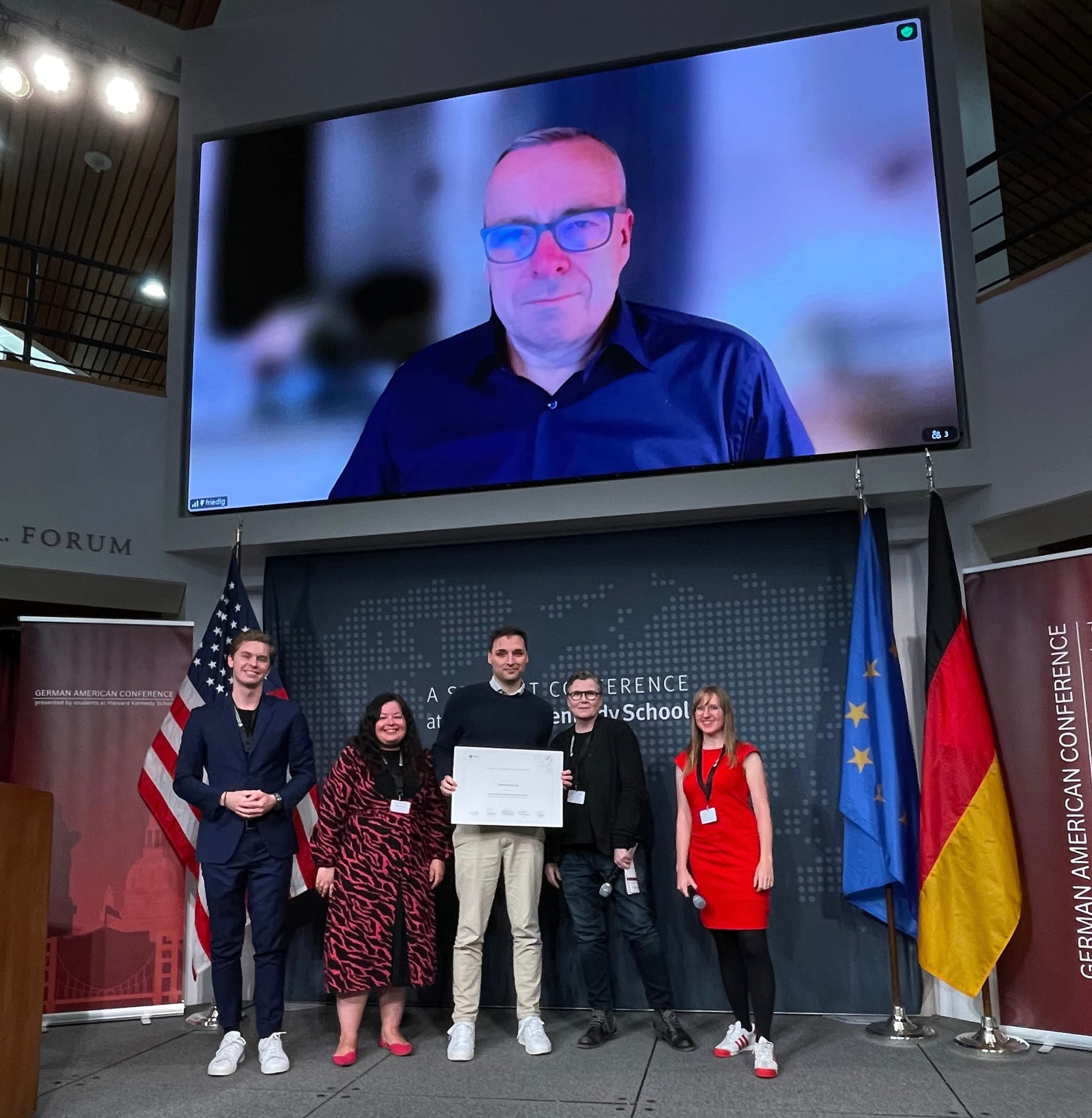 Auf der Bühne der German American Conference an der Harvard Kennedy School: fünf Personen mit einer Urkunde, darüber eine per Video zugeschaltete Person auf der Leinwand, daneben die Flaggen der USA, der EU und Deutschlands.